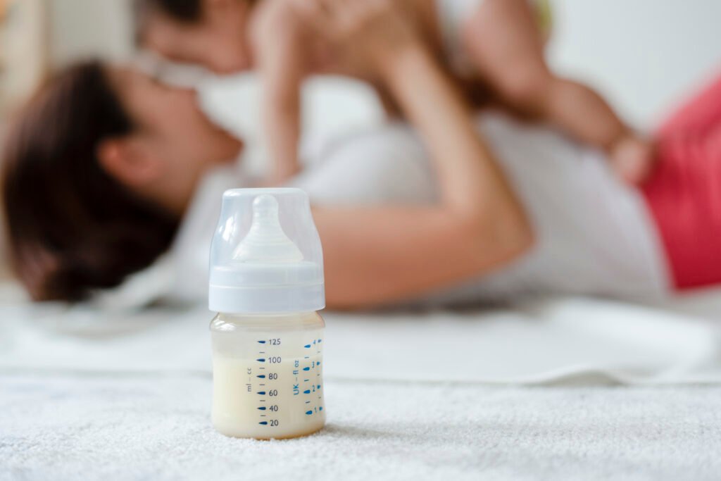 Baby bottle in focus with blurred background of mother holding baby—representing the need to stop feeding to sleep for better rest.