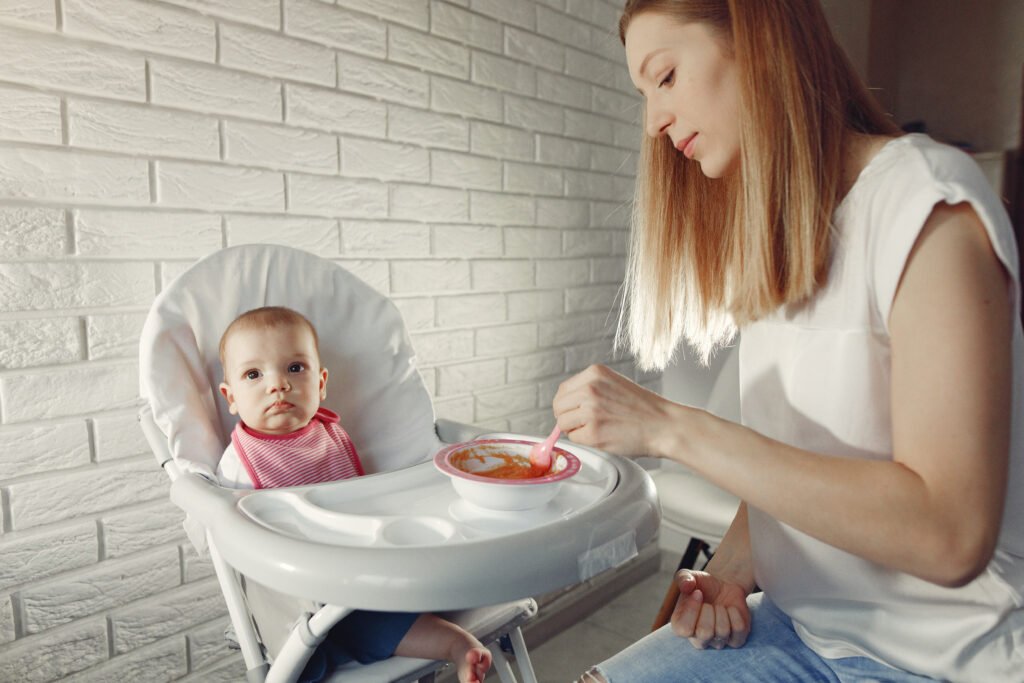 Mother introducing solids to her baby in a high chair as part of a baby-fed and spoon-fed approach.