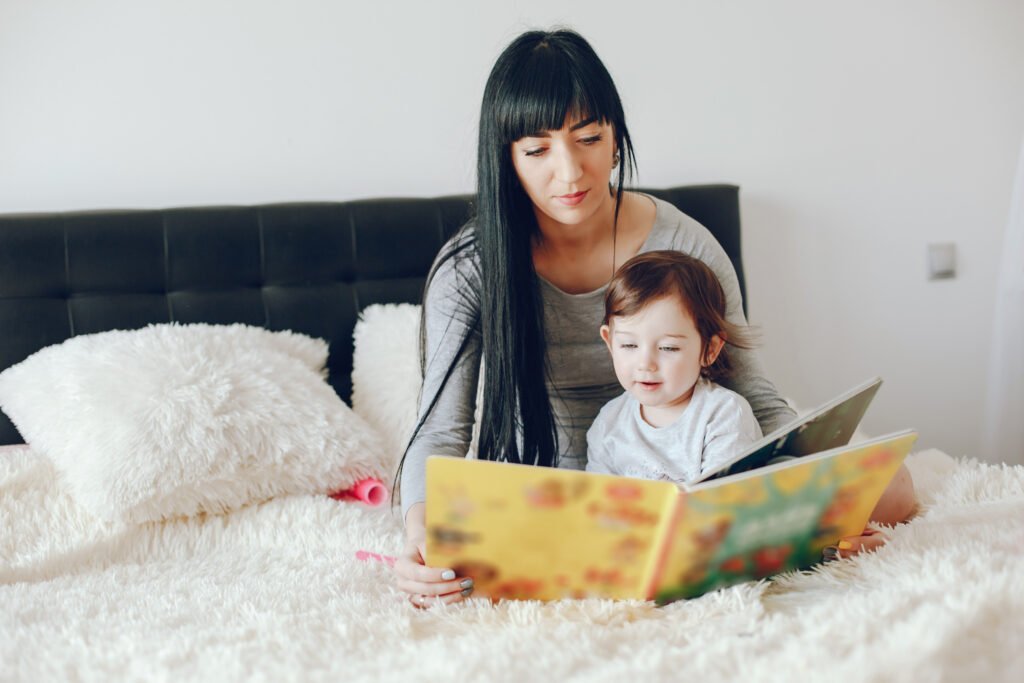 A mother and her young daughter cuddled together on a plush bed, both looking at a colorful picture book as they share a calm bedtime reading moment