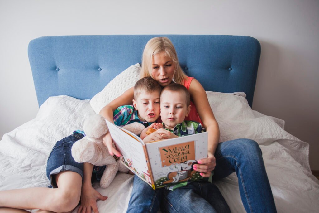 A mother in a pink top and jeans sits against a blue headboard on a white bedspread, holding an open picture book as her two young sons snuggle close—one clutching a teddy bear—while they all look at the illustrations together.